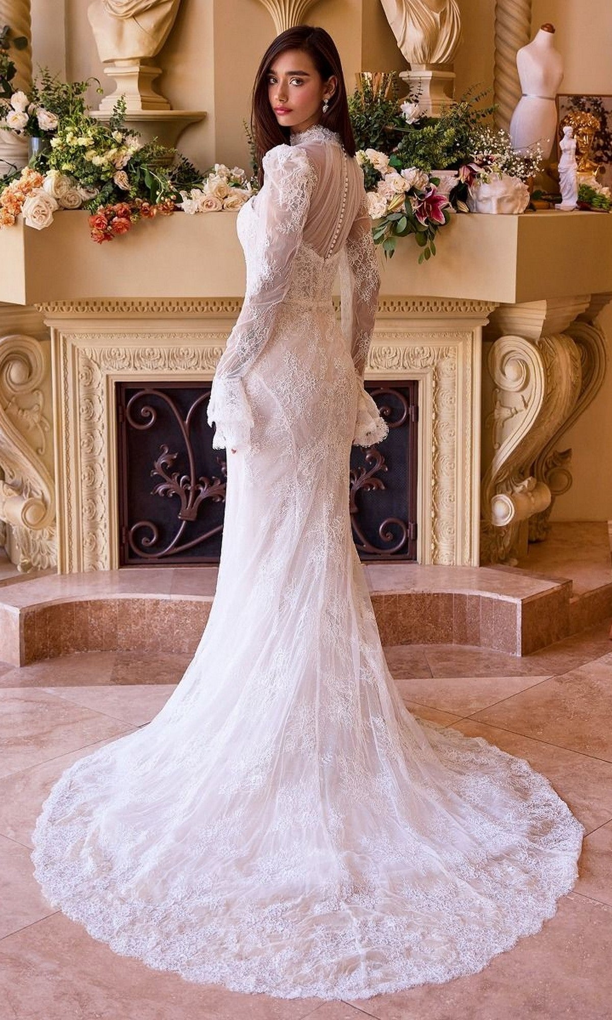 A woman in the Andrea and Leo WL042 long wedding dress with removable sleeves stands indoors, facing away, surrounded by floral arrangements and classical decor.